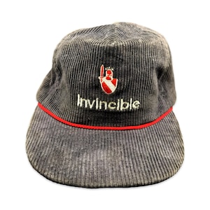 May include: A dark grey corduroy baseball cap featuring a red and white embroidered shield emblem with a sword icon above the white text Invincible. A thin red cord trim separates the crown from the flat brim of this structured headwear piece.