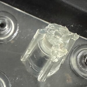 Pu&ograve; includere: Primo piano di un componente in plastica trasparente di forma irregolare, con una base cilindrica e una parte superiore parzialmente rotta. Il componente &egrave; posto su una superficie metallica con rientranze circolari.