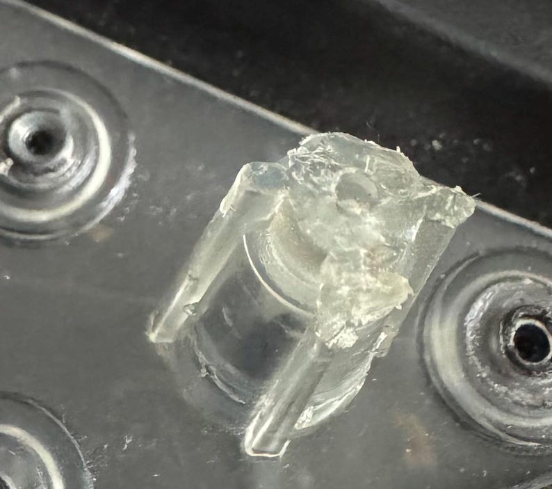 Pu&ograve; includere: Primo piano di un componente in plastica trasparente di forma irregolare, con una base cilindrica e una parte superiore parzialmente rotta. Il componente &egrave; posto su una superficie metallica con rientranze circolari.