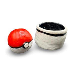 May include: A red and white ceramic Pokeball with a matching white and black container. The Pokeball is round with a white band and button. The container is cylindrical with a black interior and a black band.