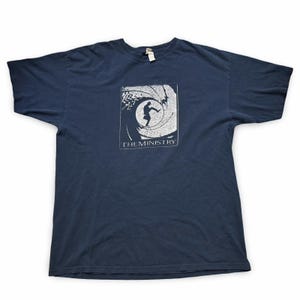 Puede incluir: Camiseta azul marino con un gráfico blanco de una persona en un diseño en espiral y el texto "THE MINISTRY". La camiseta tiene mangas cortas y cuello redondo.