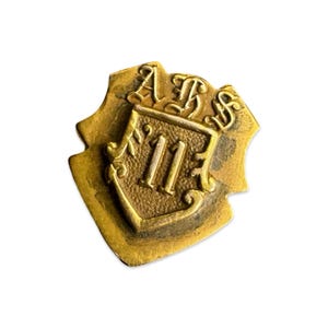 May include: A vintage gold-toned metal pin with a shield shape. The pin features the letters "ABS" at the top and the number "11" in the center. The design has ornate detailing and a slightly aged appearance.