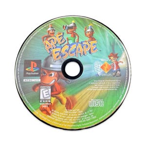 Peut inclure: Un disque de jeu PlayStation pour "Ape Escape". Le disque présente un design coloré avec le titre du jeu et des personnages, dont des singes et un garçon. Le disque porte le logo PlayStation et la classification ESRB.