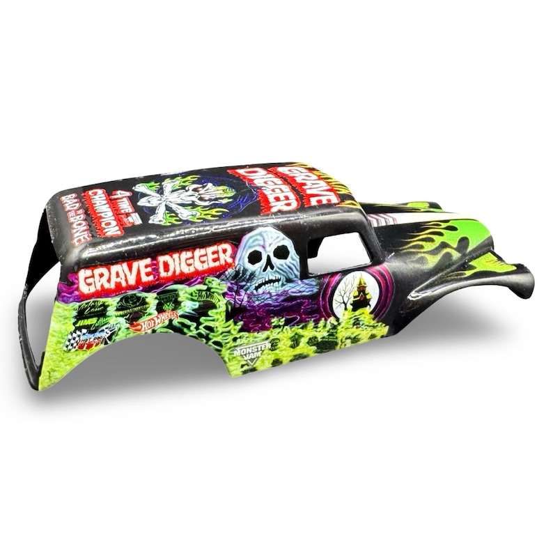 Pu&ograve; includere: Carrozzeria di monster truck Grave Digger nera e verde con grafica a teschio. Il corpo presenta le parole "Grave Digger" e "Champion" in rosso e bianco. Fiamme e altre grafiche decorano i lati.