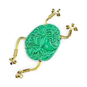 Puede incluir: Broche verde con forma de rana con un diseño floral y de pájaros tallado. El broche tiene patas y pies de alambre dorado y está sobre un fondo blanco. El cuerpo de la rana es de color verde vibrante.