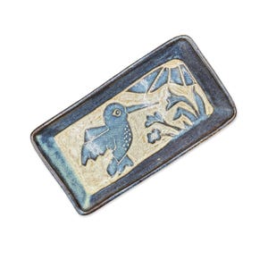 May include: A rectangular ceramic tray featuring a blue and cream glaze. The center design depicts a hummingbird in flight near stylized flowers and a sunburst pattern. The tray has a dark blue border and a textured, hand-crafted appearance.