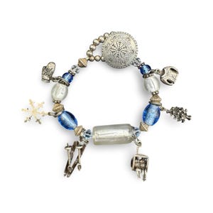Könnte beinhalten: Ein silberfarbenes Charm-Armband mit winterlichen Anhängern. Das Armband hat blaue und klare Perlen, einen Schneeflocken-Anhänger, Miniatur-Skier, einen Fäustling und einen dekorativen runden Verschluss mit geprägtem Design.