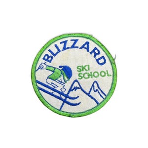 Może przedstawiać: Okrągła naszywka z zieloną obwódką i napisem "BLIZZARD SKI SCHOOL" w kolorze niebieskim i zielonym. Przedstawia narciarza w kolorze zielonym i niebieskim zjeżdżającego z góry. Tło jest białe.