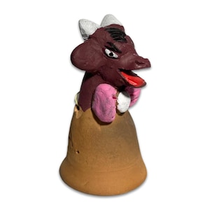 May include: A handmade ceramic figurine of a creature with a dark maroon head, white horns, and a red tongue. It has pink and white accents and is set on a brown bell-shaped base. The figurine has an expressive, slightly angry face.
