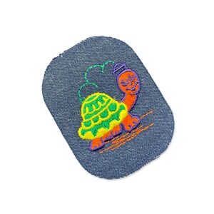 May include: A rectangular denim patch with rounded corners featuring an embroidered turtle. The turtle has an orange head and limbs, a bright green and yellow shell, and a purple hat. Green dashed lines arc above the turtle's shell.