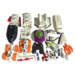 May include: A collection of toy space vehicles and accessories, including rockets, a shuttle, a rover, and a UFO. The toys are primarily white, with orange, black, red, and green accents. Includes astronauts and a control panel.