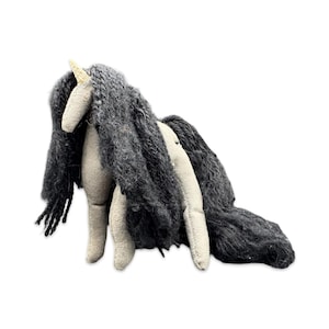May include: A handmade plush unicorn with a cream-colored body, gray mane, and a golden horn. The unicorn's long, flowing mane is made of a textured, dark gray yarn. The toy is designed to stand on its four legs.