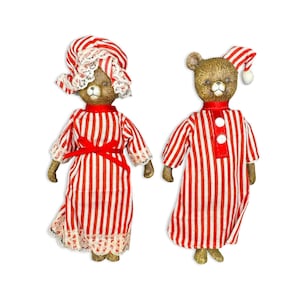 May include: Two brown bear figurines dressed in red and white striped pyjamas. The left bear wears a matching nightcap with white lace trim and a red ribbon belt. The right bear wears a red and white striped nightcap with a white pom-pom.