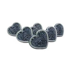 Op de afbeelding: Zes hartvormige decoratieve objecten in een gedempte blauwe tint, elk met een wit bloemmotief. De objecten zijn in een halve cirkel gerangschikt, met de harten naar de kijker gericht. De achtergrond is effen wit.