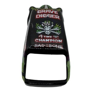 Pu&ograve; includere: Giocattolo di camion mostro Grave Digger nero e verde. Il giocattolo presenta il logo Grave Digger, un design con teschio e ossa incrociate e la scritta "4 Time Champion". Il giocattolo ha una forma rettangolare con la parte anteriore aperta.