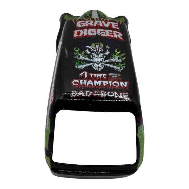 Pu&ograve; includere: Giocattolo di camion mostro Grave Digger nero e verde. Il giocattolo presenta il logo Grave Digger, un design con teschio e ossa incrociate e la scritta "4 Time Champion". Il giocattolo ha una forma rettangolare con la parte anteriore aperta.