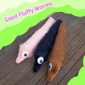May include: Three plush, fluffy worms in pink, black, and brown, each with large, googly eyes. The text "Giant Fluffy Worms" is displayed above the worms. The worms are lying on a wooden surface.