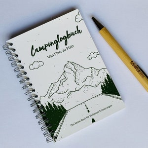 Könnte beinhalten: Ein weißes Spiralblock mit einer schwarz-weißen Illustration einer Bergkette und einer Straße. Das Notizbuch sagt "Campinglogbuch Von Platz zu Platz" und "Das kleine Buch für bleibende Erinnerungen". Ein Holzstift mit dem Wort "Camperstyle" liegt neben dem Notizbuch.