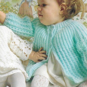 May include: A baby in a light blue knitted cardigan with ribbed detailing. The baby is also wearing a white dress and lace patterned tights. The cardigan has long sleeves and a round neckline. The baby is sitting on a white surface.