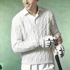 May include: A white cable knit sweater with a v-neck. The sweater is worn by a person holding a cricket bat and wearing white cricket gloves.