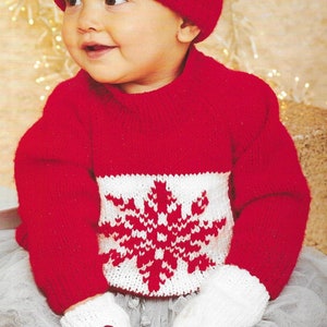 May include: A red knitted baby sweater with a white snowflake design. The sweater has a red knitted hat with a white snowflake design and a white pom-pom on top. The baby is wearing a white tulle skirt.