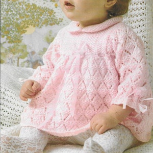 May include: A baby girl wearing a pink knitted dress with a white lace trim and a pink ribbon tied at the waist. The dress has a diamond pattern and is crocheted.