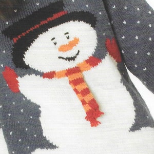 Pu&ograve; includere: Un maglione di lana bianco con un disegno di pupazzo di neve. Il pupazzo di neve indossa un cappello nero, una sciarpa rosso e arancione e guanti rossi. Il maglione ha uno sfondo grigio con pois bianchi.