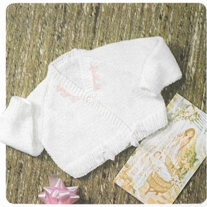 Op de afbeelding: Een wit gebreid babyvest met knoopsluiting en roze bies. Het vest ligt op een bruine ondergrond met een kaart en een roze strik.