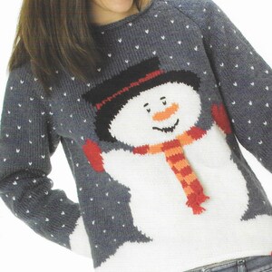 Pu&ograve; includere: Un maglione di lana grigio con un disegno di pupazzo di neve bianco. Il pupazzo di neve ha un cappello nero, un naso arancione e una sciarpa rossa. Il maglione ha un motivo a fiocco di neve bianco.