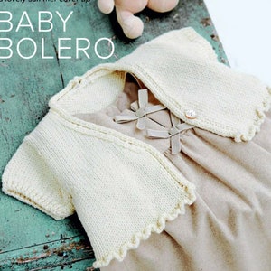 May include: A white knitted baby bolero with a scalloped edge. The bolero is worn over a brown dress with a bow detail.
