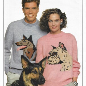 May include: Two people wearing sweaters with dog illustrations. The man is wearing a light blue sweater with a German Shepherd illustration. The woman is wearing a pink sweater with a Boxer illustration. A German Shepherd dog is standing in front of them.