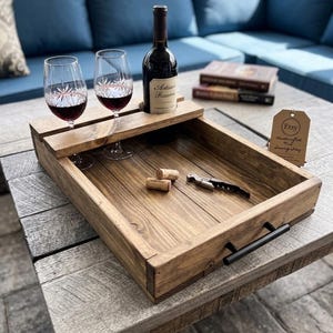May include: A rectangular wooden serving tray with a dark brown finish and black metal handles. The tray features a built-in rack for two wine glasses and a wine bottle. Inside the tray are two corks and a metal corkscrew. A brown tag reads Etsy Handcrafted Wine Serving Tray.