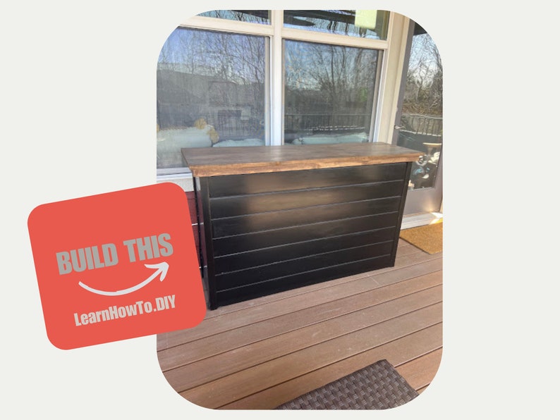 May include: A black wooden bar with a wood countertop, on a wooden deck. The bar has a horizontal slat design. The text "BUILD THIS" and "LearnHowTo.DIY" is on a red background.