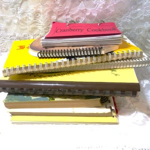 May include: A stack of spiral bound notebooks with yellow, brown, and red covers. The top notebook has the title "Cranberry Cookbook" on the cover.