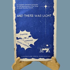 May include: A vintage blue Christmas program cover with the text "AND THERE WAS LIGHT." The cover features a star, a manger scene, and the words "Inspiring," "Different," and "Christ exalting." It is displayed on a wooden stand.