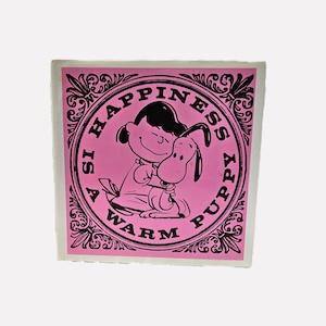 May include: A pink square featuring a black and white illustration of a girl hugging a dog. The text "Happiness is a Warm Puppy" is in a circular frame. The design is on a white background.