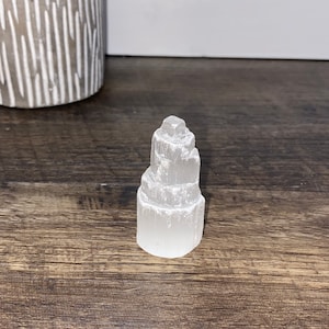 May include: A white selenite crystal tower with a layered, stacked design. The crystal is smooth and polished, with a natural, earthy look.
