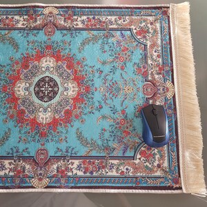 Könnte beinhalten: Ein rechteckiges Mauspad mit einem leuchtend türkisfarbenen Hintergrund, einem detaillierten Blumenmedaillonmuster in Rot, Weiß und Dunkelblau sowie aufwendigen Bordüren. Das Pad hat cremefarbene Fransen an der rechten Kante und eine schwarze Maus.