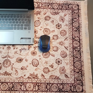 May include: A laptop and a blue and black wireless mouse rest on a cream-colored, floral-patterned rug. The rug has a brown border and fringe along one edge. The laptop is silver and has a Lenovo sticker.