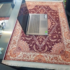 May include: A laptop computer rests on a rectangular, ornate rug. The rug features a burgundy background with a cream-colored border and intricate floral patterns. The rug has a fringe along the edges.