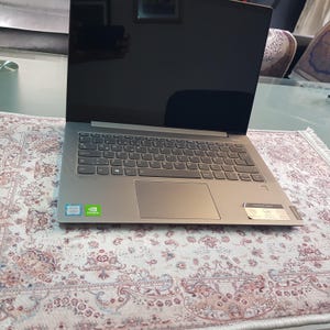 May include: A silver laptop with a black screen, open on a floral patterned rug. The keyboard is visible, with a touchpad below. The laptop has Intel and NVIDIA stickers. The model name is visible on the bottom right.