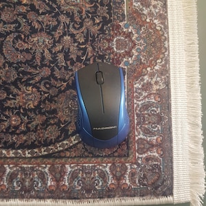 May include: A wireless computer mouse with a blue and black design, resting on a patterned rug. The mouse has a scroll wheel and the brand name "HADRON" is visible. The rug features a detailed floral pattern with a cream-colored fringe.