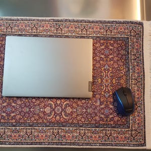 May include: A silver Lenovo laptop and a blue and black wireless mouse rest on a decorative rectangular rug. The rug features a burgundy background with an intricate floral pattern and a cream-colored fringe.