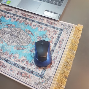 May include: A blue and black wireless computer mouse rests on a decorative mousepad with a traditional rug design in shades of blue, cream, and pink. The mousepad has a fringed edge, and a laptop is partially visible in the background.