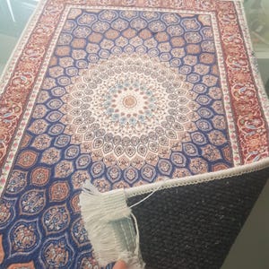 May include: A rectangular rug with an intricate floral pattern in shades of blue, cream, and rust. The design features a central medallion surrounded by repeating motifs. The rug has a fringed edge and a dark, textured backing.