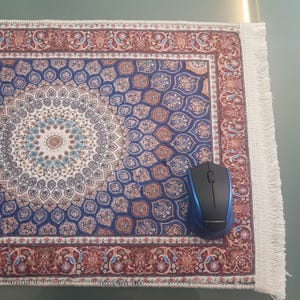 May include: A decorative mousepad with a Persian rug design in shades of blue, brown, and cream. The mousepad has a detailed floral pattern and a fringed edge. A blue and black wireless mouse rests on the pad.