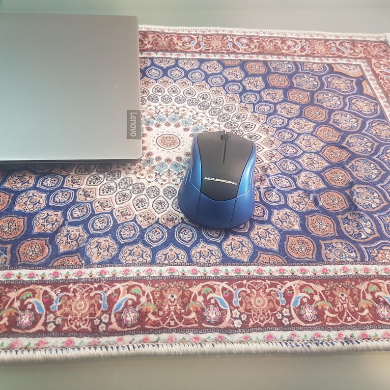 May include: A blue and black computer mouse sits on a patterned desk mat with a laptop. The mat features a detailed design with blue, red, and cream colors, resembling a miniature rug. The laptop is silver with the Lenovo logo.