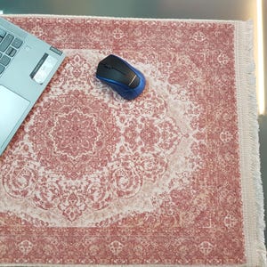 May include: A decorative mousepad with a traditional rug design in shades of red and cream. A blue and black wireless mouse and a laptop are on the mousepad. The mousepad has a fringed edge.