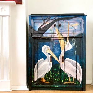 May include: A dark blue painted cabinet with two doors and a single drawer. The doors feature a hand-painted scene of two white pelicans in a swamp setting with green reeds and a blue sky.