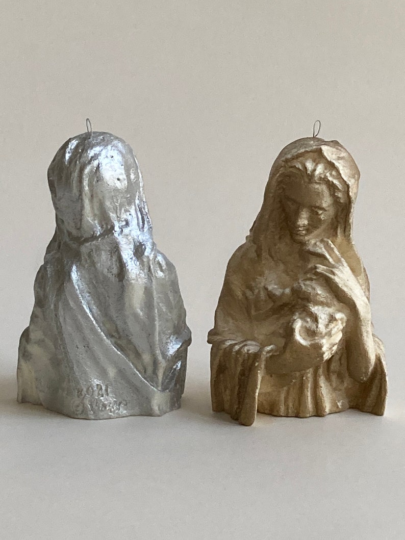 May include: Two small, decorative figurines of a woman holding a child. The figurine on the left is silver, and the figurine on the right is gold.